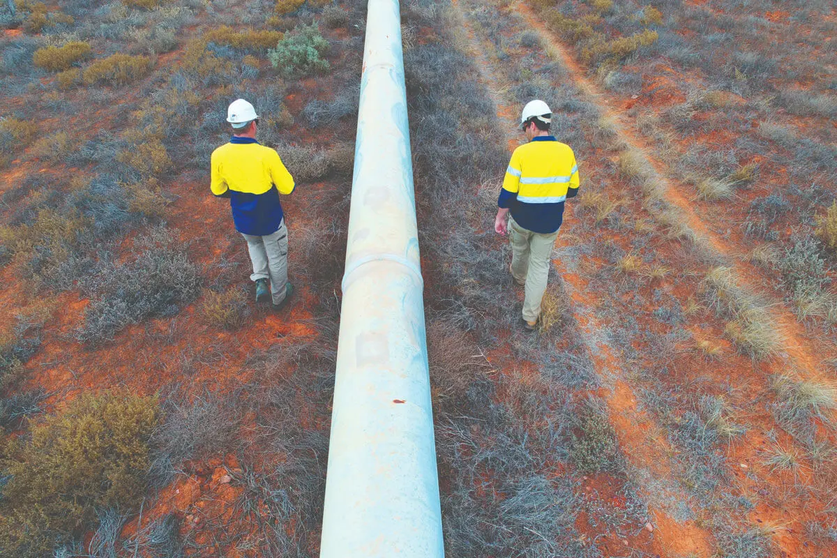 Two workers in yellow and blue safety jackets and white helmets walk beside a large pipeline in a dry, red-soil landscape with sparse vegetation, demonstrating attention to personal care and workplace safety.