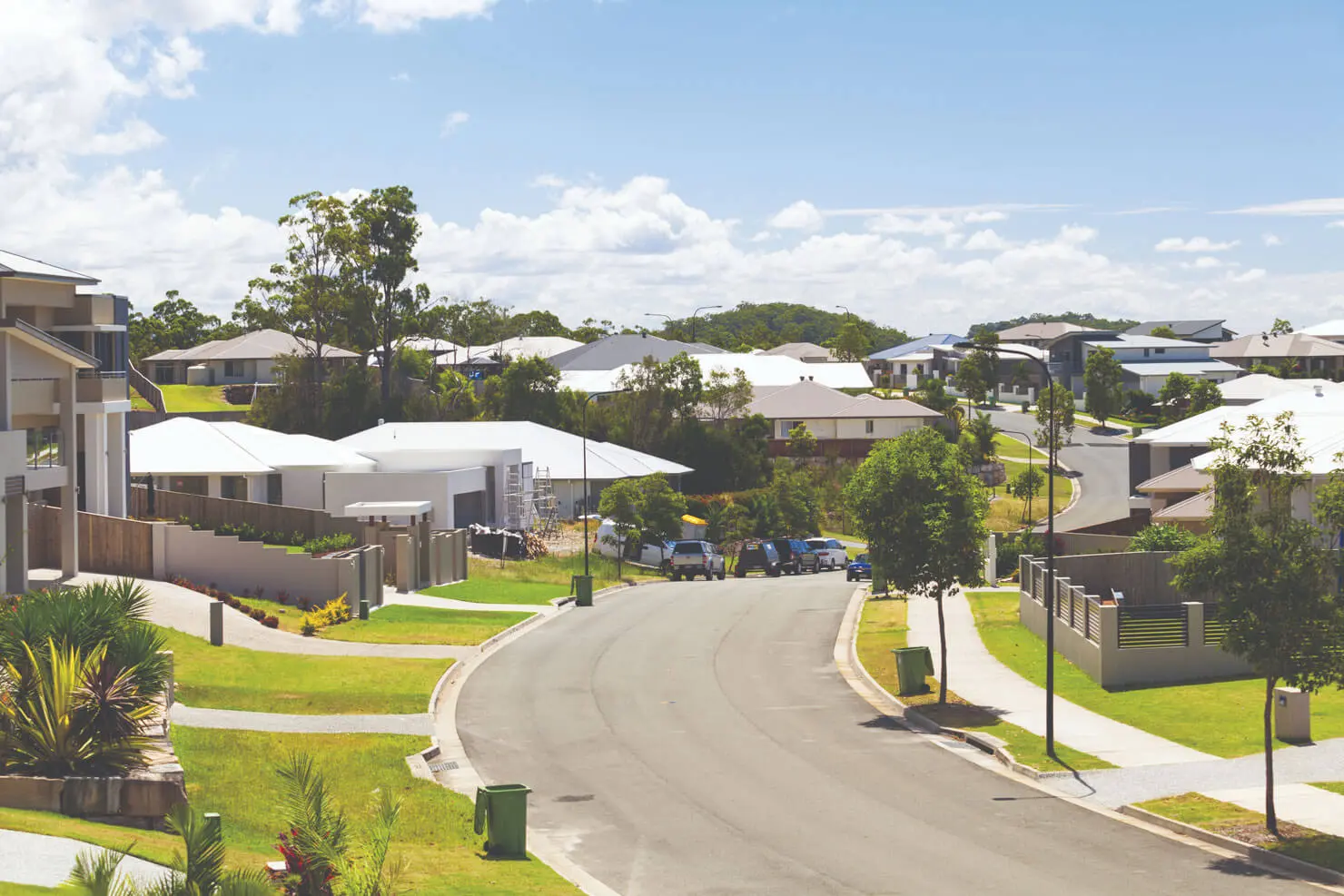A quiet suburban street curves through a modern neighborhood with neatly manicured lawns, trees, parked cars, and houses featuring light-colored roofs under a blue sky with scattered clouds.