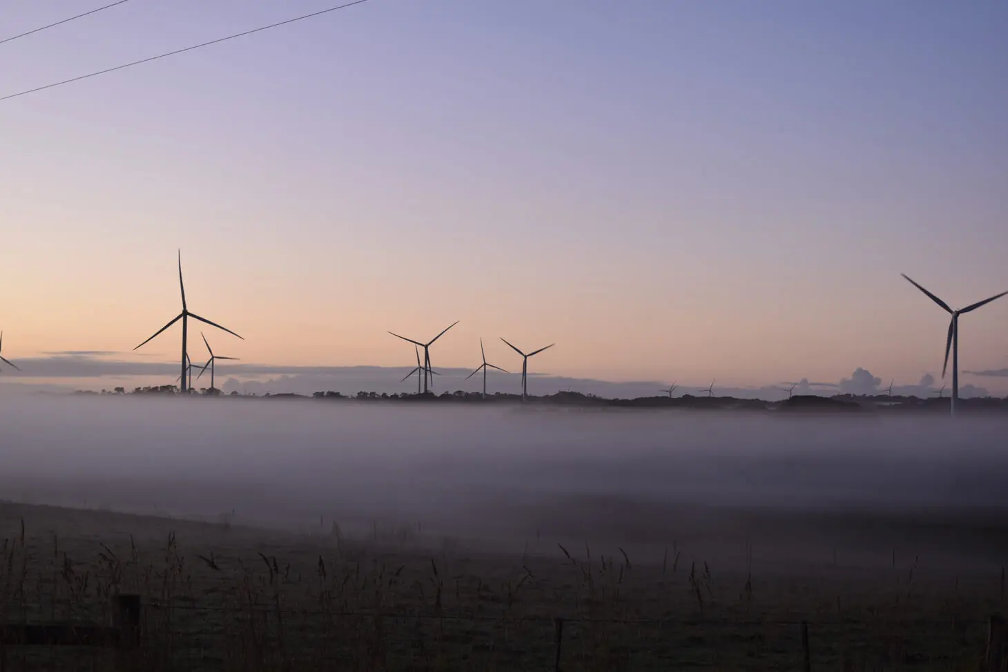 Wind turbines stand in a misty field at dawn, with soft sunlight and a pale sky in the background. The landscape is partially obscured by low fog, creating a serene and tranquil scene.