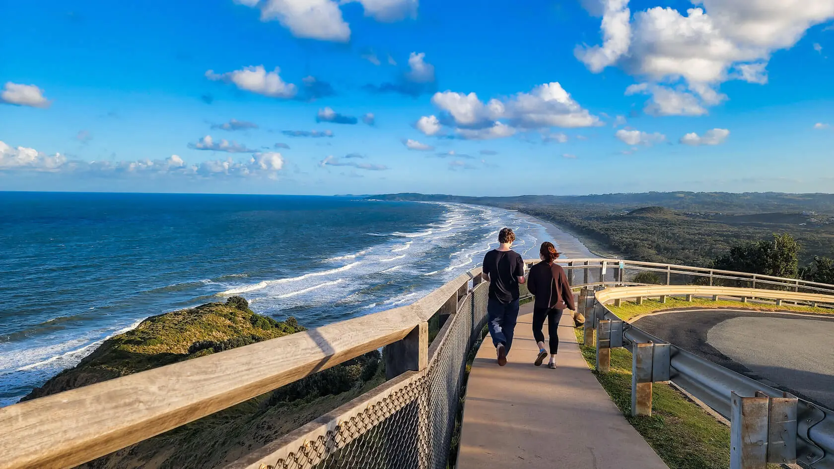 Two people walk on a paved path along a wooden fence overlooking a scenic coastline. This accessible walkway, suitable for NDIS participants, offers breathtaking views of waves, cliffs, and a blue sky dotted with clouds.