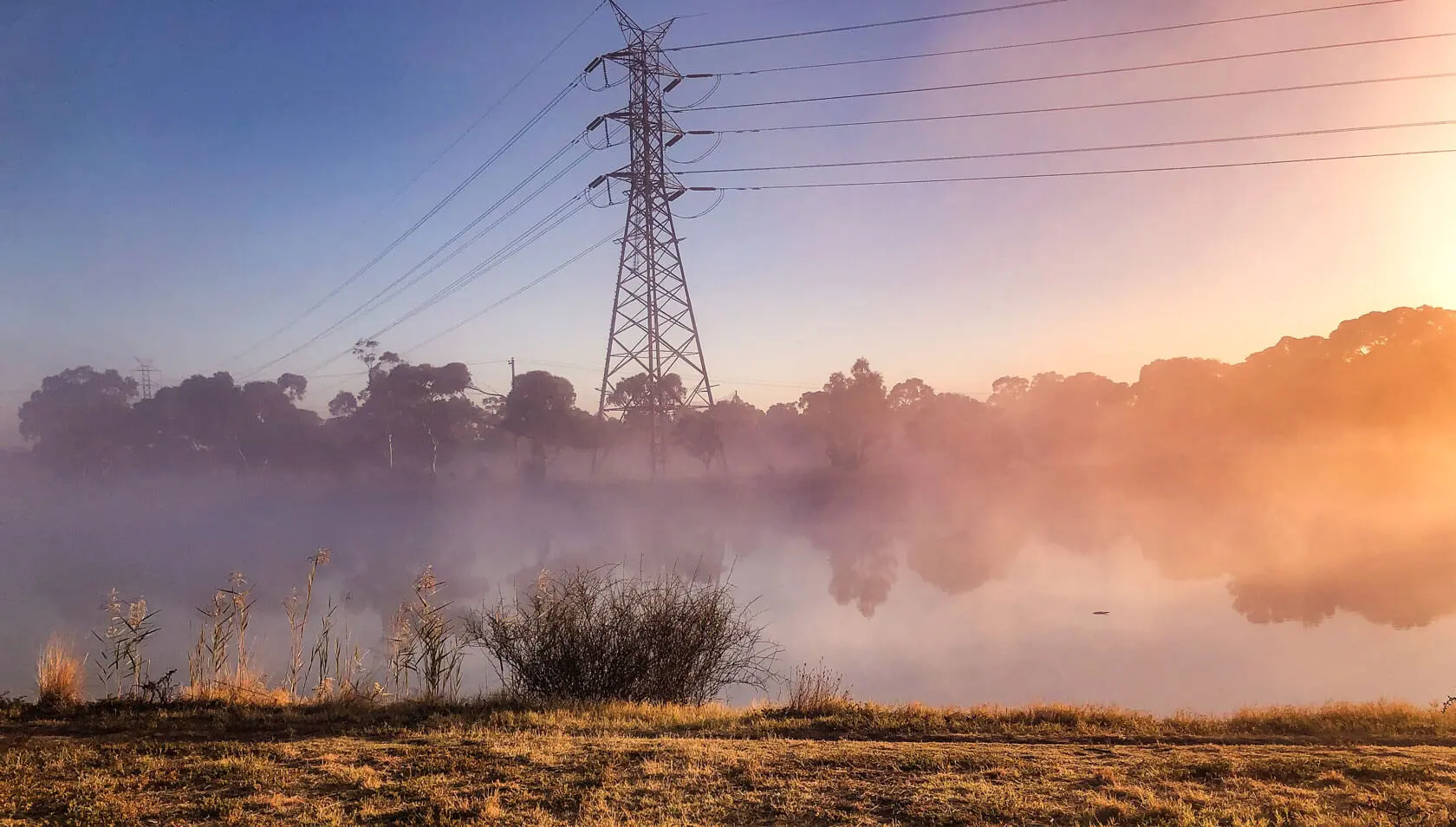 A power line tower stands near a calm body of water, with trees and mist in the background. Soft sunlight creates a pink and orange haze over the foggy landscape, symbolizing quiet strength—much like support provided through the NDIS.